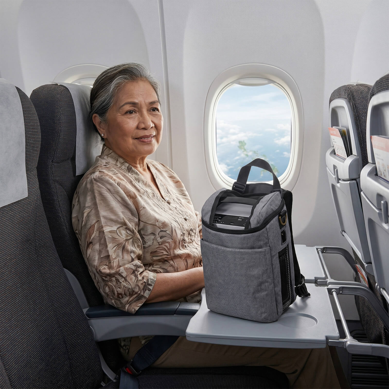An elderly woman traveling on a plane with a VP-1 Portable Oxygen Concentrator in a shoulder bag.